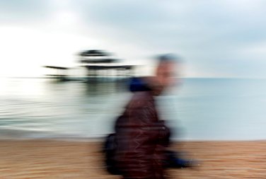 A motion blurred photo with Brighton West Pier in the background and a male student with his camera walking past in the frontground. Photo by Eva Kalpadaki.