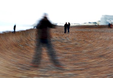 Circular motion blur photograph by Eva Kalpadaki showing a male student standing in a pose A on the pebbles in Brighton seafront with three other people in the back ground.