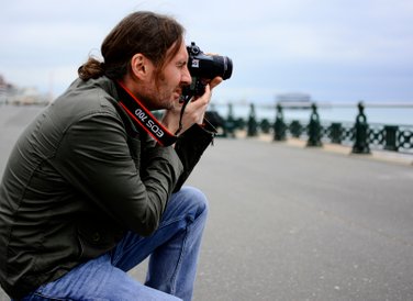 A male student is kneeling on the ground in Hove promenade holding his camera on his eye facing the sea with West Pier in shallow focus in the background. Photo by Eva Kalpadaki. 