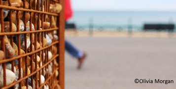 View of a Brighton seafront grid full of pebbles cage with the leg and a back of a man behind it and the sea and a bench in the blurry background. Photo by Olivia Morgan.
