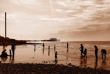 Sepia view of Brighton West Pier and i360 from Hove with silhouetted people and kids playing in the shallow waters and the sandy beach. Photo by Eva Kalpadaki.