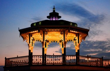 Side view of yellow lit Brighton Bandstand in the night in blue background sky. Photo by Eva Kalpadaki.