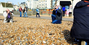 Three male students taking photos on the Brighton seafront while kneeling on the pebbles. Photo by Eva Kalpadaki.