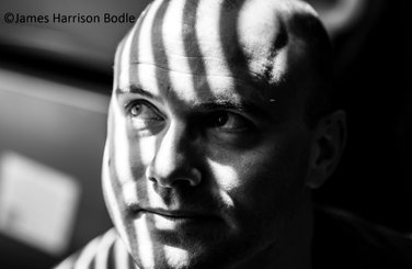 Black and white photo by James Harrison Bodle showing only the face of a bald man with stripes of light projected on his forehead while he is looking upwards to the right.