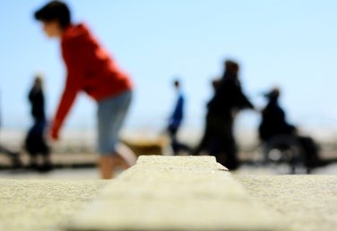 Out of focus photograph by Eva Kalpadaki taken from a low angle in front of the concrete sitting places on Brighton seafront. In the background fuzzy figures are seen walking past in the blue background of the sky with a boy in red hoodie and a woman pushing another in a wheelchair as the most prominent ones.